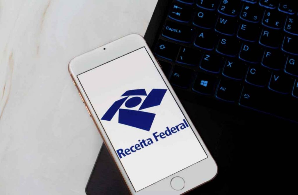 Celular com o logotipo da Receita Federal na tela, apoiado ao lado de um notebook