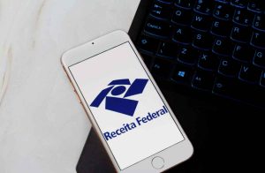 Celular com o logotipo da Receita Federal na tela, apoiado ao lado de um notebook